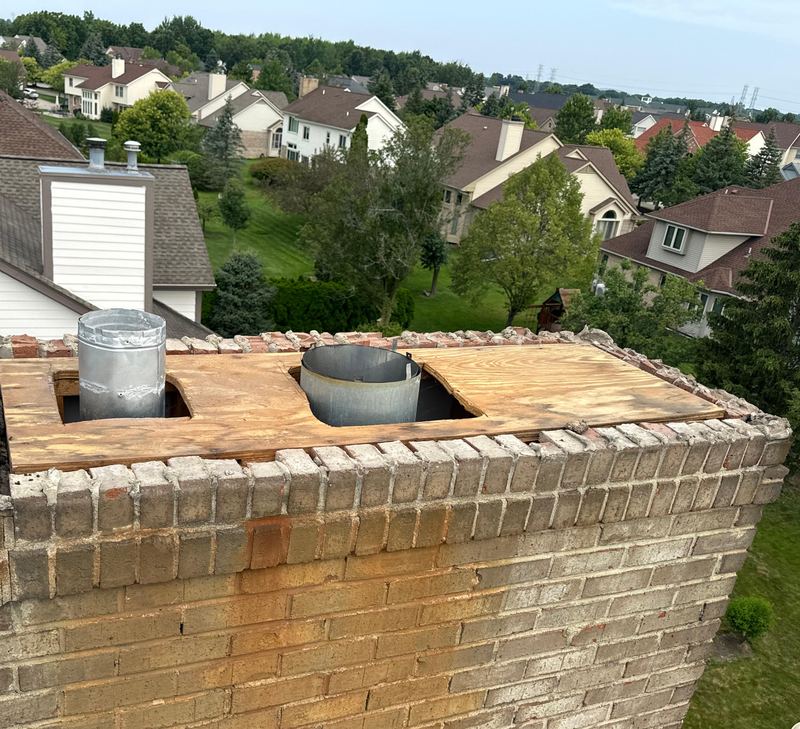 Tan brick chimney top with the crown gone, plywood over the opening around two metal flues